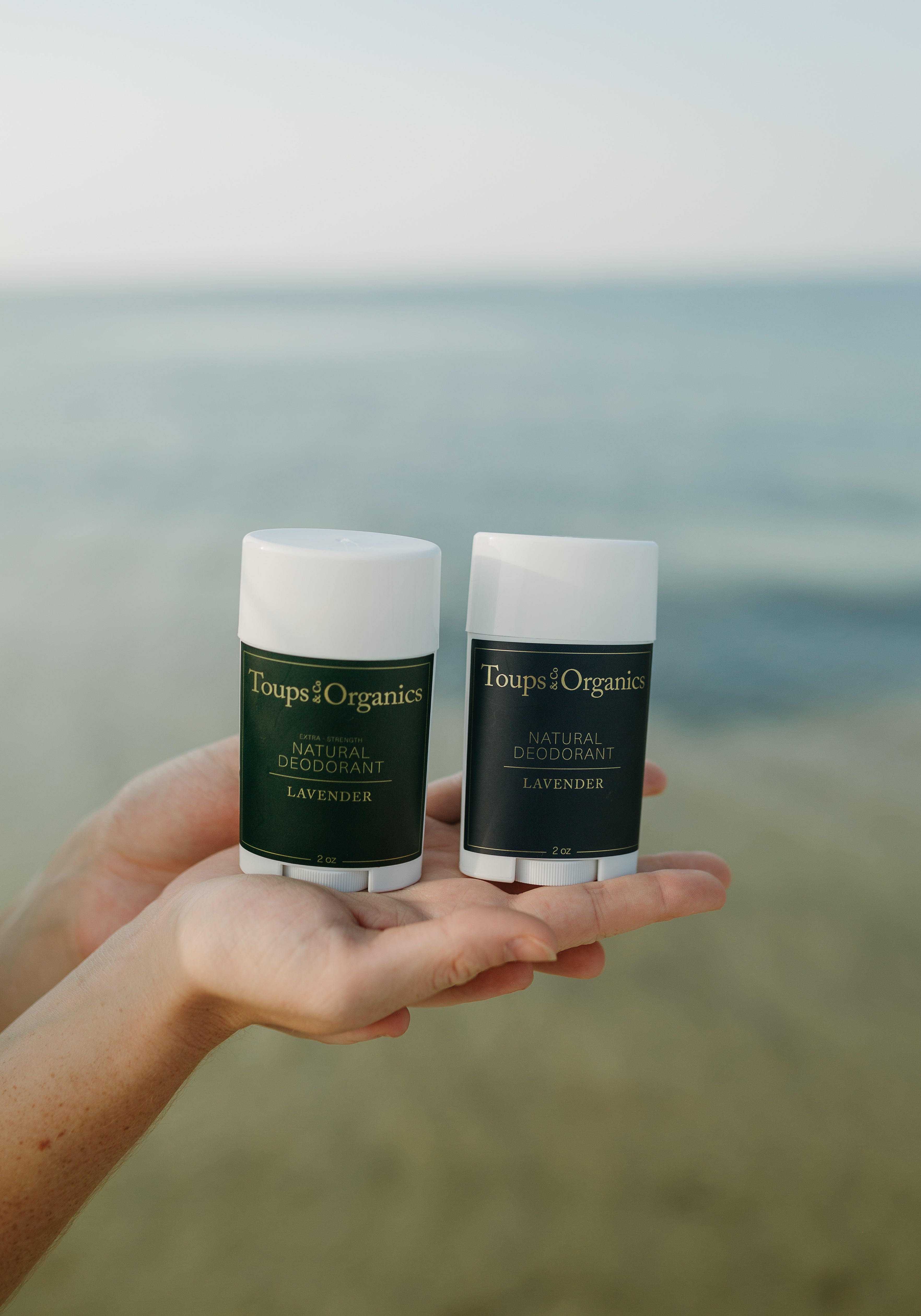 woman-holding-two-tubes-of-natural-deodorant-against-ocean-background