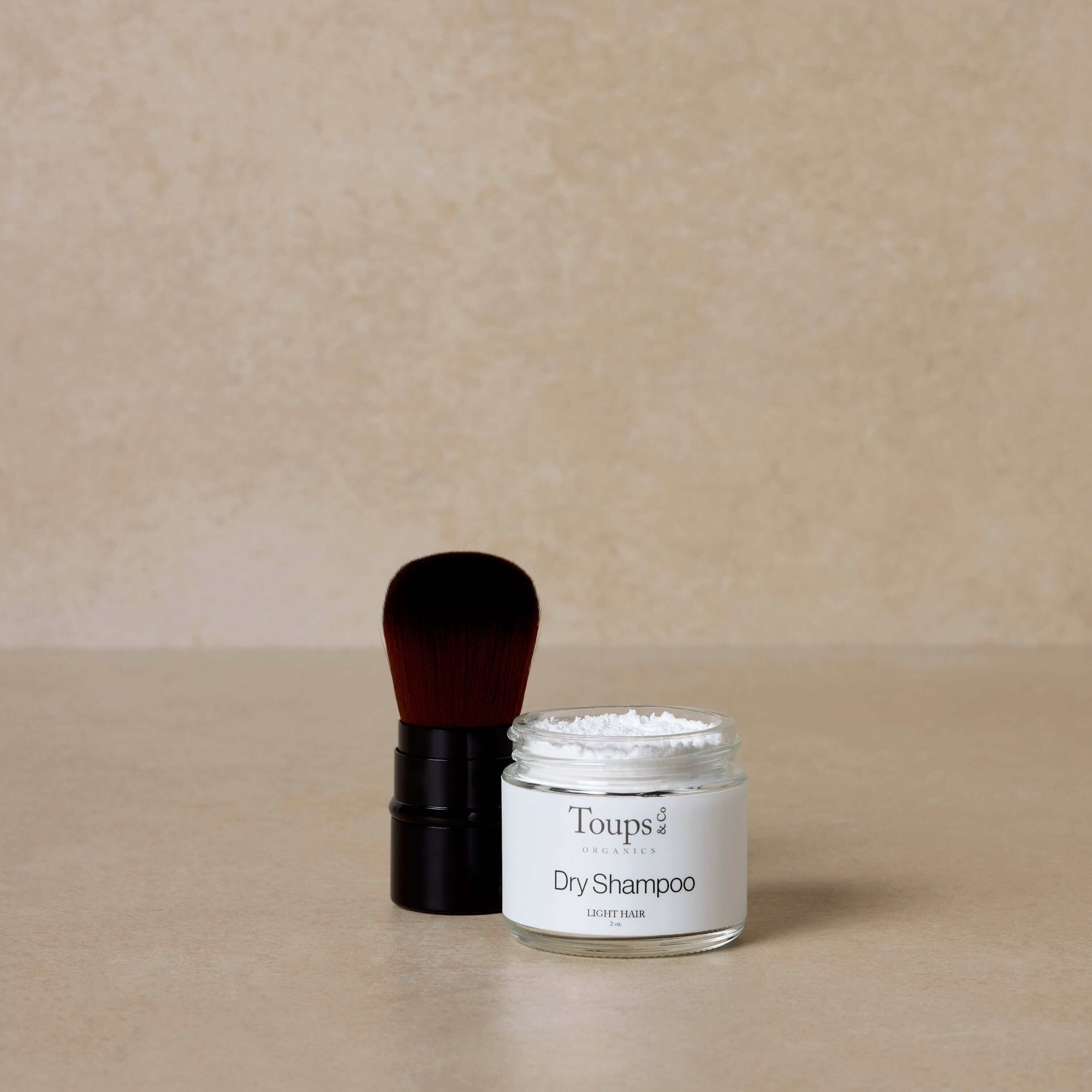 A small Dry Shampoo + Applicator Set from Toups and Co Organics sits beside a makeup brush on a beige surface, ideal for volumizing hair with this easy-to-use bundle set against a neutral background.
