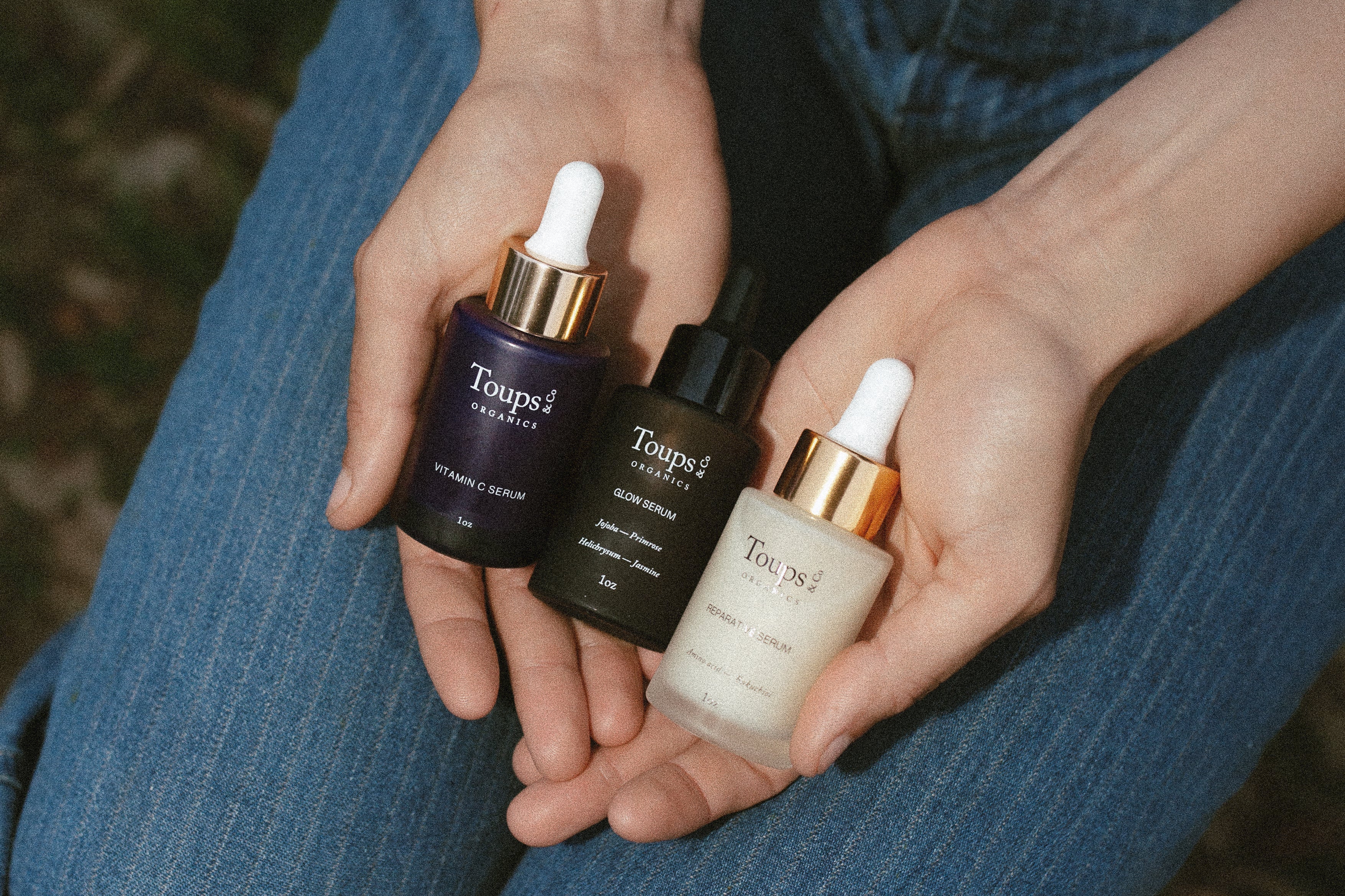 Two hands holding three Toups & Co. serums for natural skincare, including Vitamin C Serum, Glow Serum, and Reparative Serum