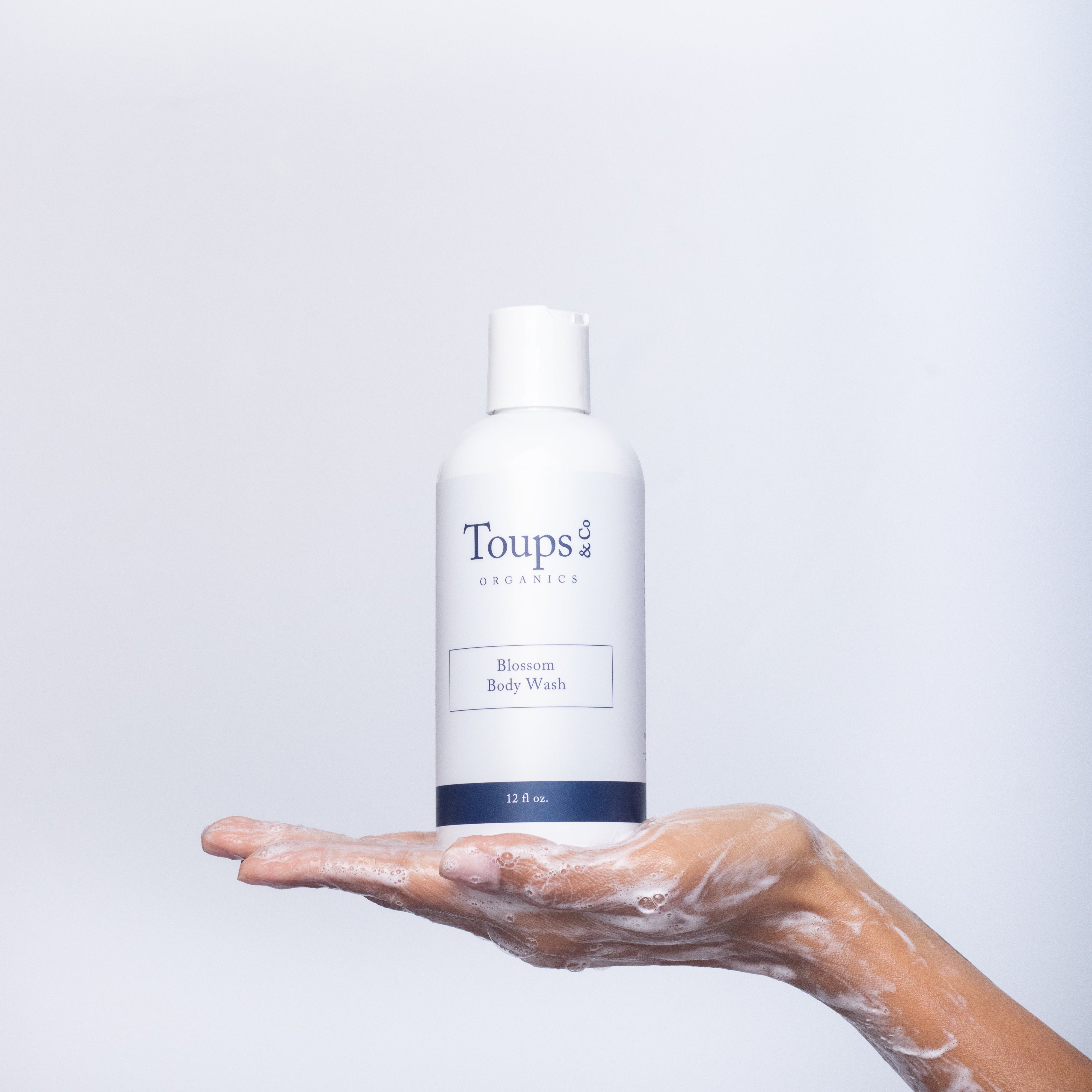 A soapy hand holds a bottle of Body Wash by Toups and Co Organics against a plain white background.