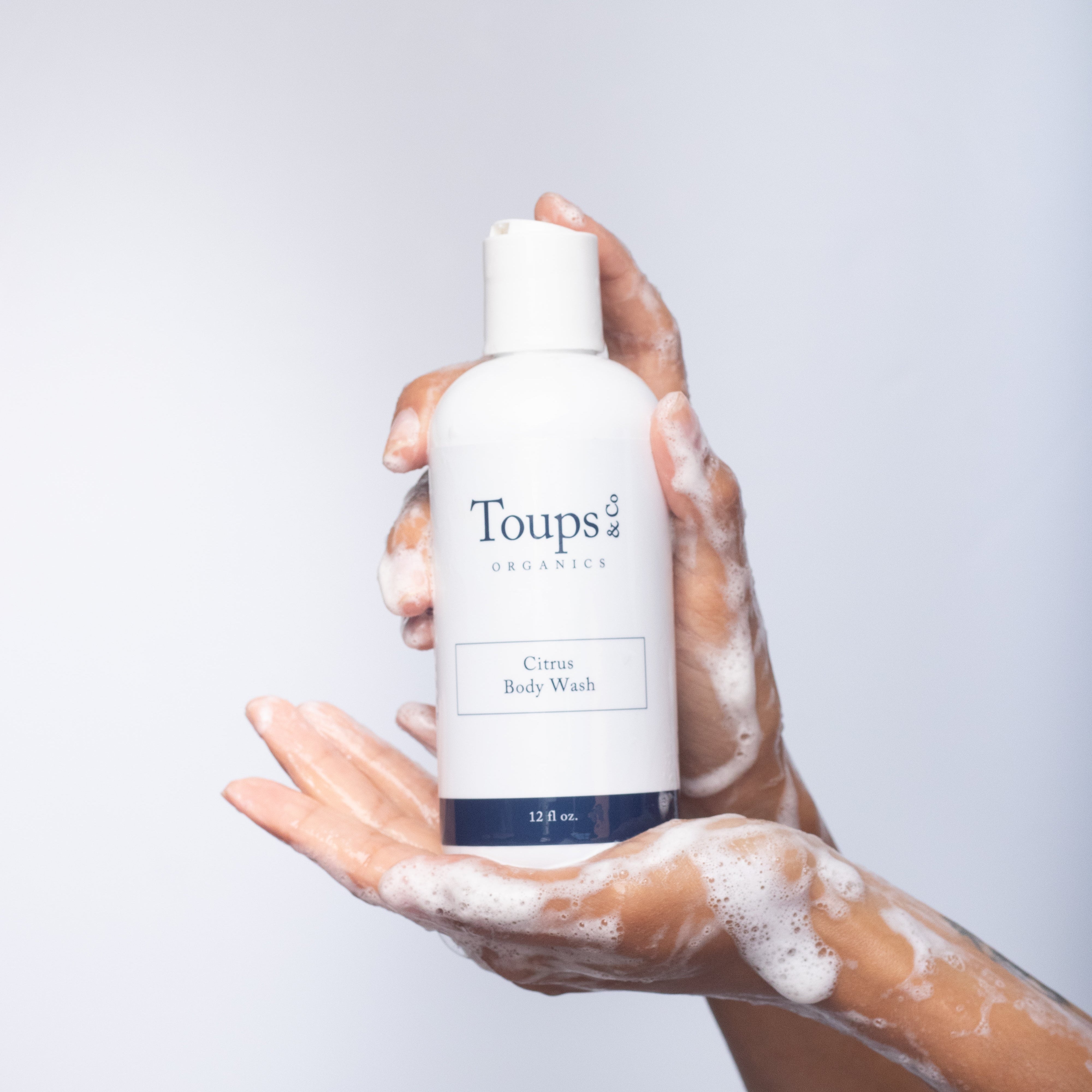 A soapy hand holds a bottle of Toups and Co Organics Body Wash against a plain white background.