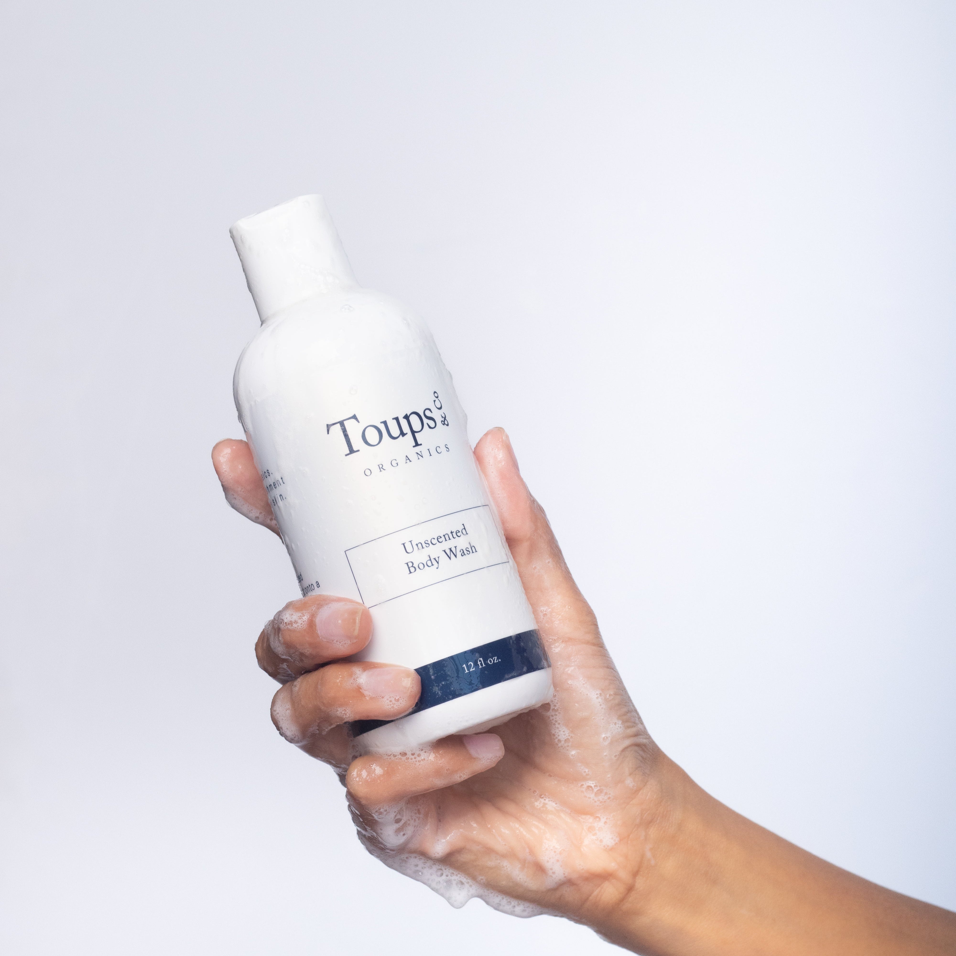 A soapy hand holds a bottle of Toups and Co Organics Body Wash against a plain white background.
