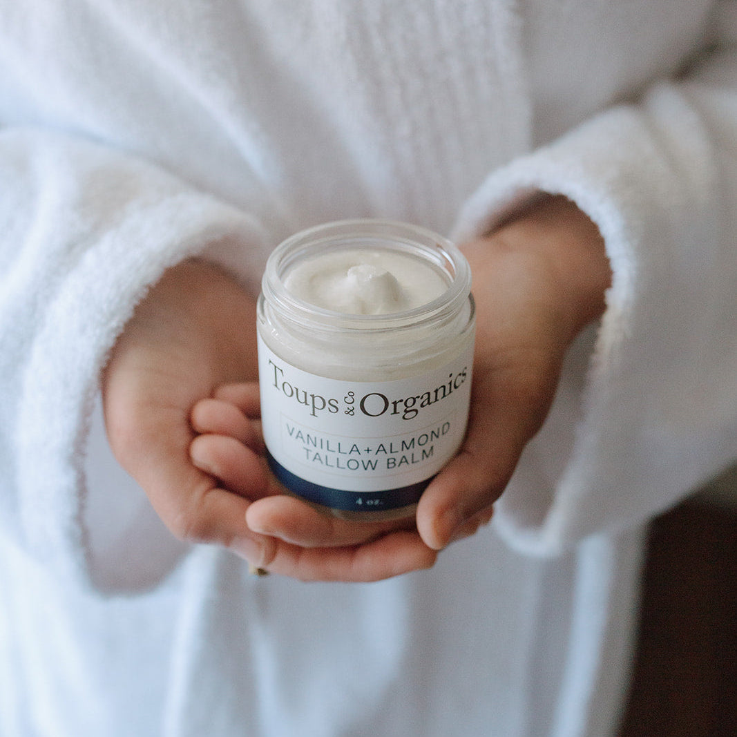 Clad in a white robe, an individual holds an open jar of Toups and Co Organics Tallow Balm - Vanilla + Almond, displaying its rich cream—a luxurious natural moisturizer crafted from nourishing, grass-fed tallow.