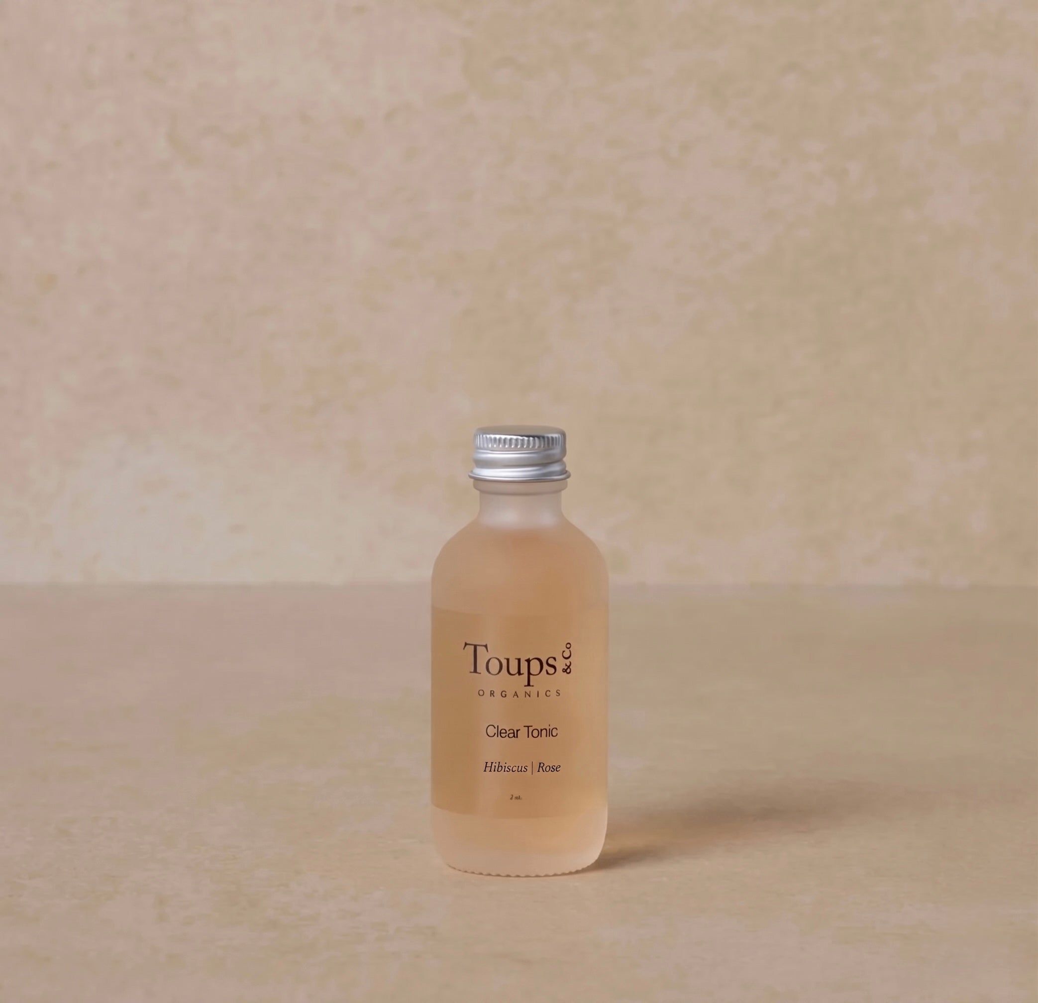 A small clear bottle labeled Toups and Co Organics Facial Tonic rests on a beige surface, blending with the matching neutral background.