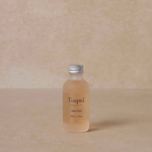 A small clear bottle labeled Toups and Co Organics Facial Tonic rests on a beige surface, blending with the matching neutral background.