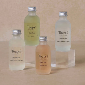 Four small Toups and Co Organics Facial Tonic bottles labeled Calm, Hydrate, Brighten, and Clear are arranged on a beige surface—some elevated on clear acrylic blocks—showcasing this luxurious organic skincare collection.