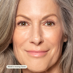 Close-up of an older woman with gray hair and smooth skin, wearing Toups and Co Organics Liquid Foundation in Oyster for a natural glow. Text reads, Model is wearing Oyster.