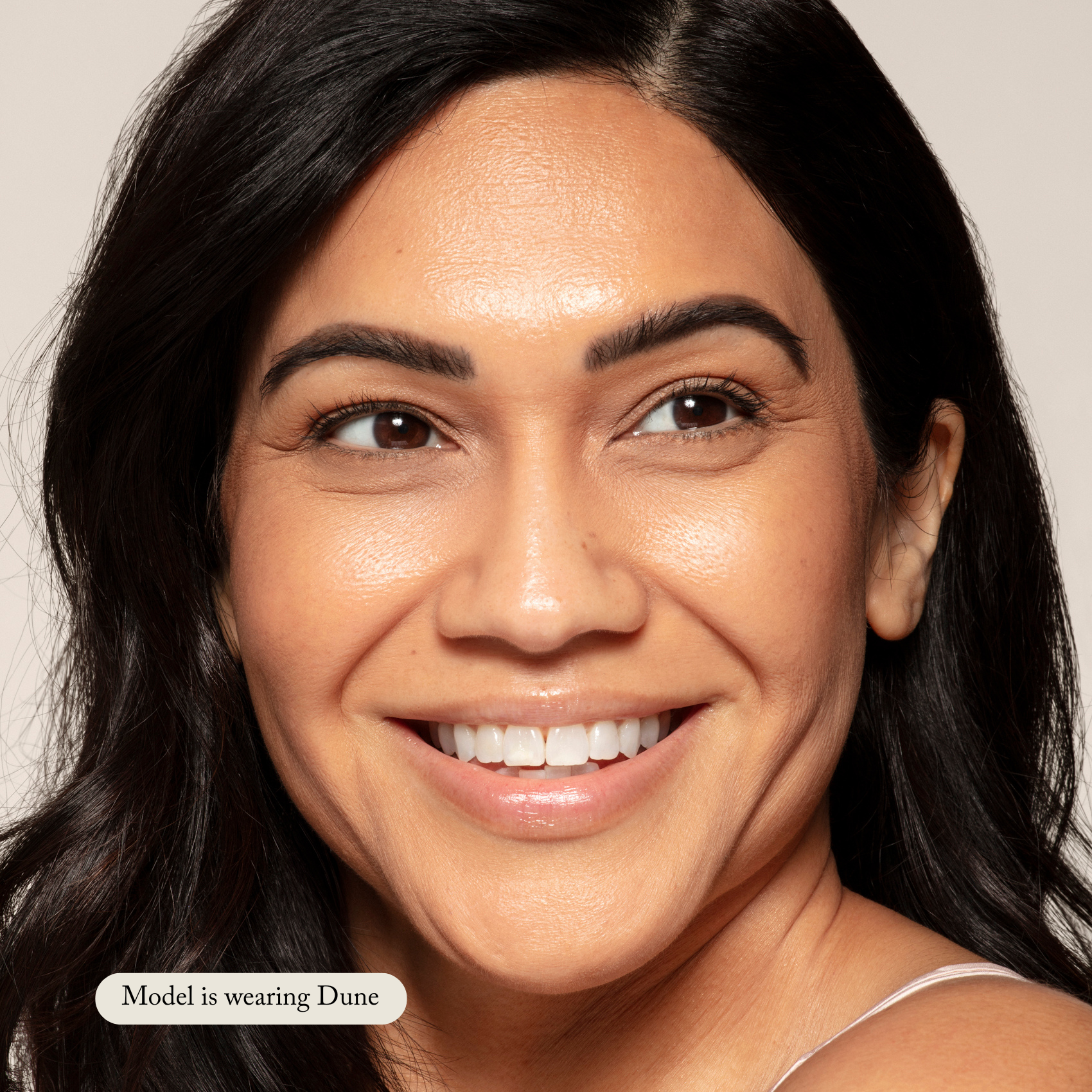 A woman with medium skin tone smiles, showing even Toups and Co Organics Liquid Foundation application for a natural glow. Text reads, Model is wearing Dune.