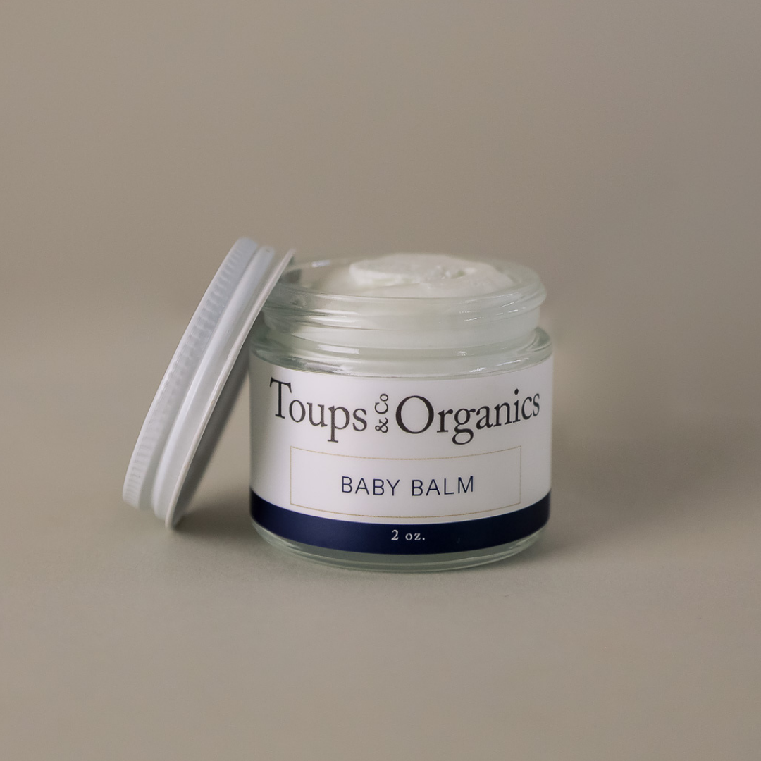A 2-ounce glass jar of Toups and Co Organics Baby Balm with the lid off reveals soothing white balm inside—ideal for easing diaper rash and eczema.