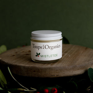 A small jar of Toups and Co Organics Mistletoe Balm sits on a wooden surface with greenery and berries, making it a perfect Christmas gift against a festive green background.