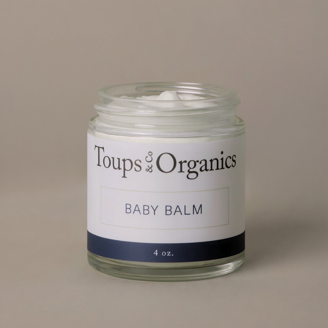 A glass jar labeled Toups and Co Organics Baby Balm, containing white balm for diaper rash or eczema relief, is set against a plain beige background.