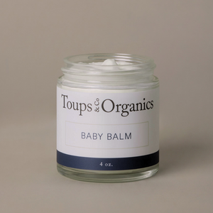 A glass jar labeled Toups and Co Organics Baby Balm, containing white balm for diaper rash or eczema relief, is set against a plain beige background.
