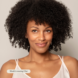A woman with natural curly hair and a light peach camisole smiles slightly, looking off-camera, her natural glow enhanced. Text reads: Model is wearing Toups and Co Organics Liquid Foundation in Chai.