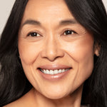 A woman with long dark hair, clear skin, and a natural smile looks slightly right, highlighting her sun-kissed glow created with Toups and Co Organics Bronzer.