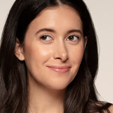 A woman with long dark hair and clear skin smiles slightly while looking off camera, her radiant complexion enhanced by Toups and Co Organics Bronzer for a sun-kissed glow, set against a plain light background.
