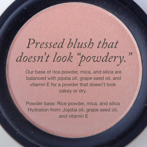 A round compact of peachy-pink pressed blush, overlaid with text describing the product as non-powdery and listing ingredients including rice powder, oils, and vitamin E.