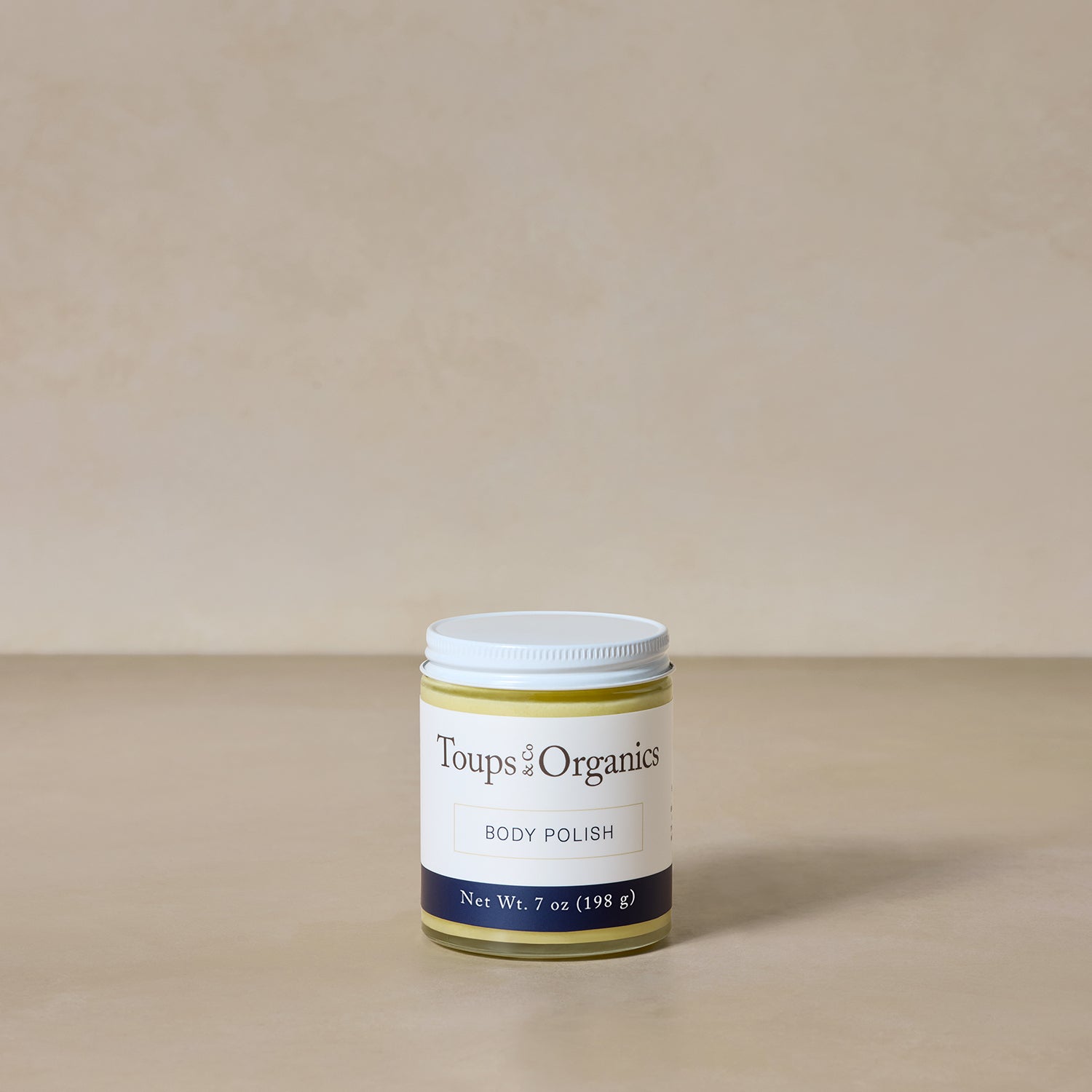 A jar of Toups and Co Organics Body Polish (7 oz/198 g) rests on a neutral-tone surface against a beige background.