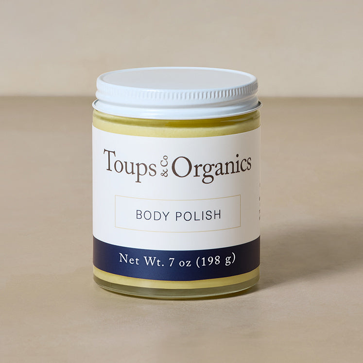 A 7 oz (198 g) glass jar of Toups and Co Organics Body Polish with a white lid is shown on a neutral beige surface.