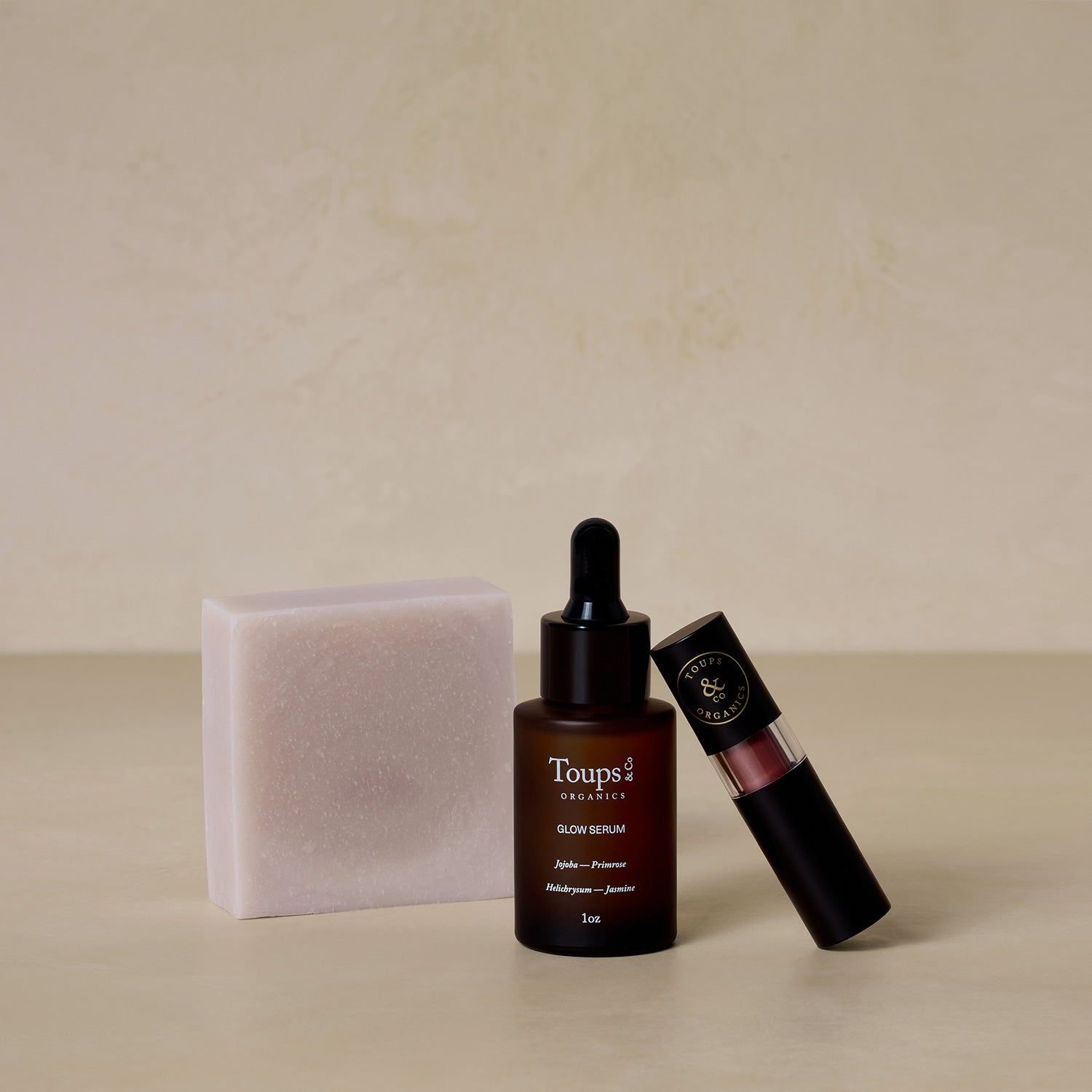 The Toups and Co Organics Blushing Bridal Set, featuring a soap bar, a brown glass Glow Serum bottle with dropper, and a tube of lip balm, is displayed on a beige surface.