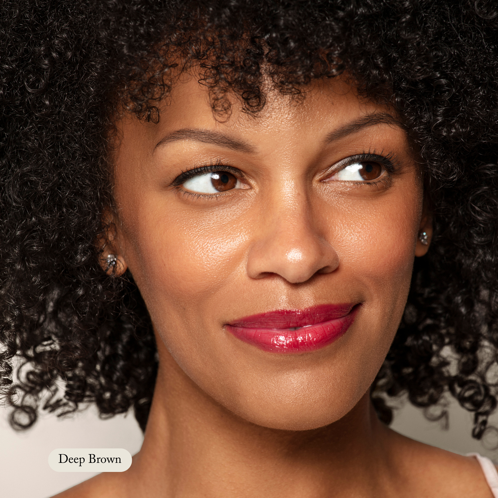 A person with curly dark hair, in glossy red lipstick, smiles softly. A label reads Deep Brown—Toups and Co Organics Brow Pencil, ideal for smooth, buttery application and perfect eyebrow color.