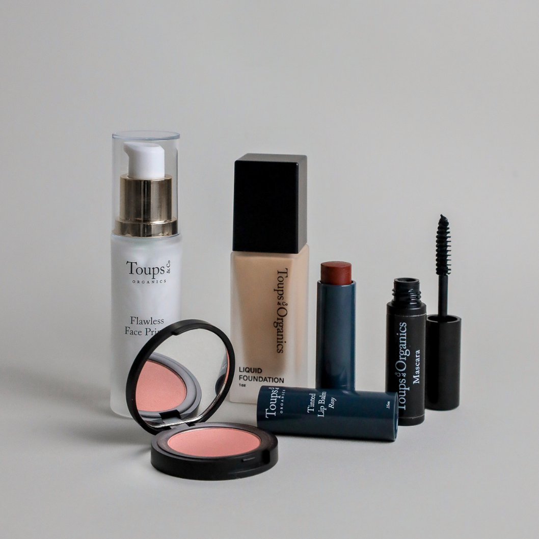 Five products from the Toups and Co Organics Complete Makeup Set—face primer, liquid foundation, blush tinted lip balm, and mascara with an open wand—are arranged on a flat surface.