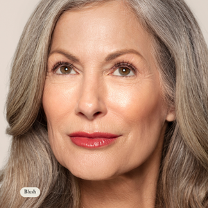 Close-up of an older woman with long gray hair and red lipstick, looking up and slightly right, showcasing a natural glow from Toups and Co Organics Cream Cheek Tint against a neutral background.