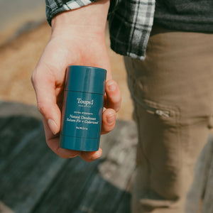 A person holding a blue stick of Toups and Co Organics Men’s Non Toxic Essentials deodorant outdoors.