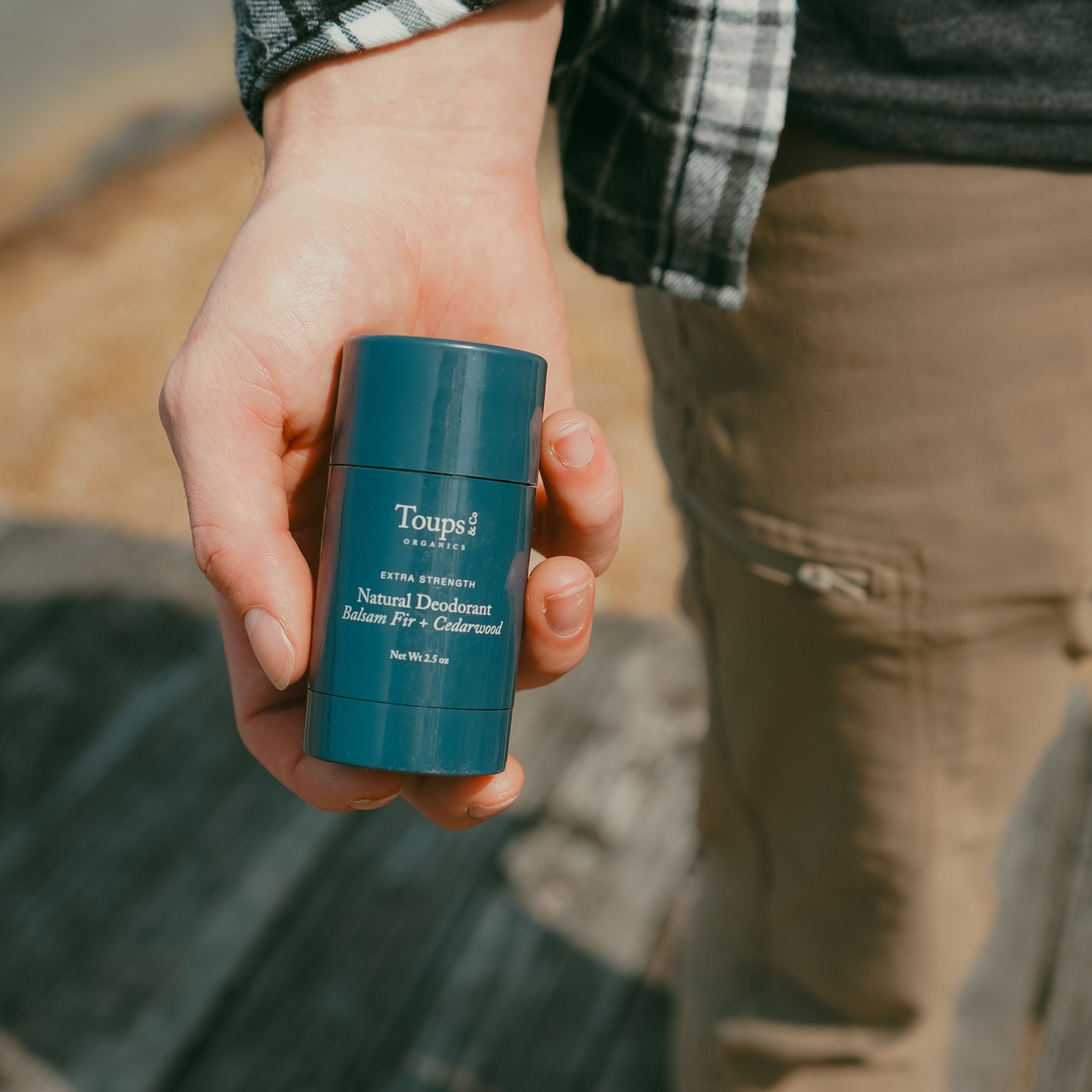 A person holding a blue stick of Toups and Co Organics Men’s Non Toxic Essentials deodorant outdoors.