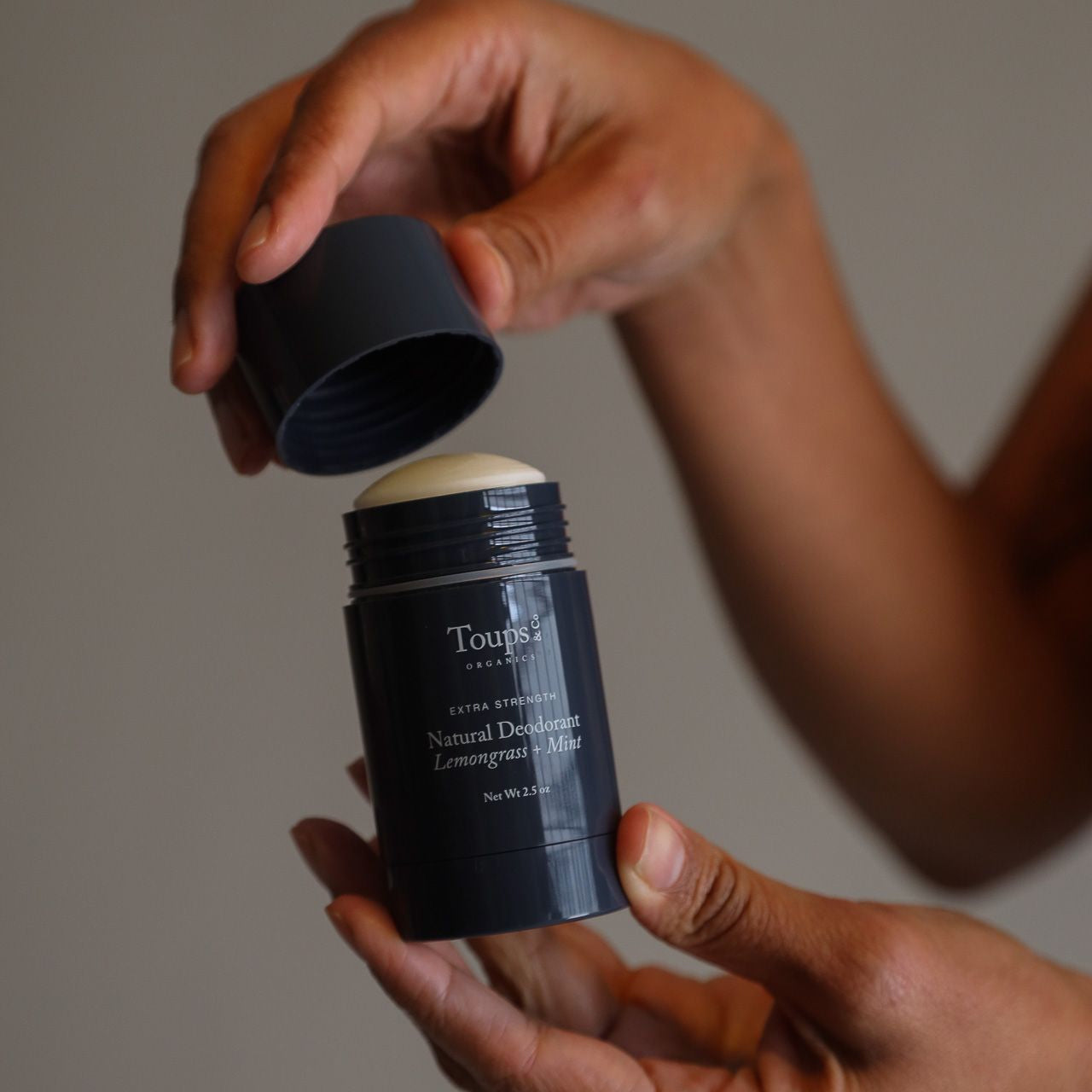 Hands opening a dark gray stick of Toups Organics Natural Deodorant in Lemongrass + Mint.