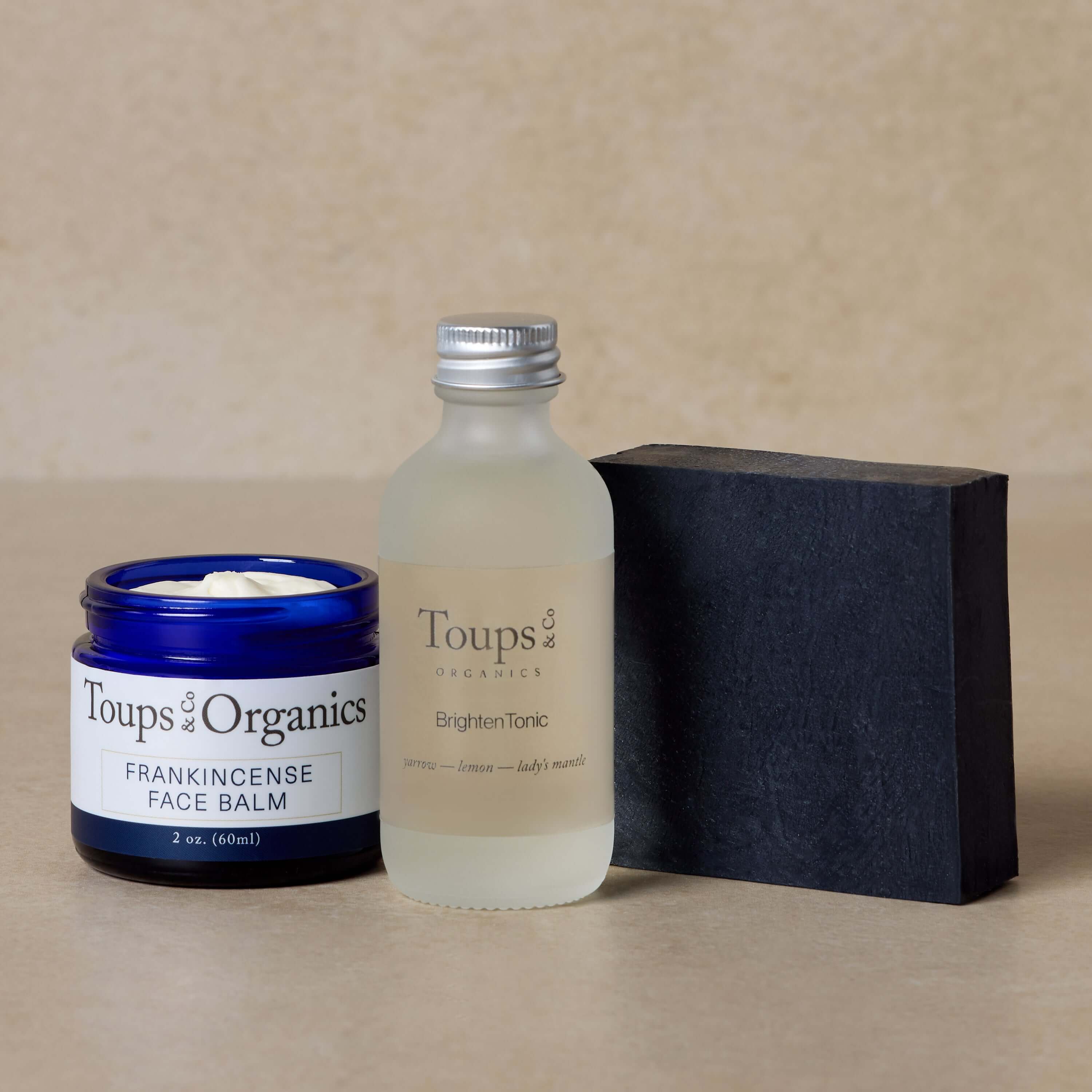 The Toups and Co Organics Essential Facial Set includes a blue jar of frankincense tallow balm, a clear bottle of brighten tonic for radiant, clear skin, and a rectangular black activated charcoal face soap bar, all displayed on a neutral background.