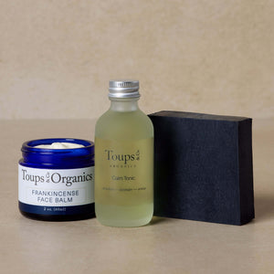 The Essential Facial Set by Toups and Co Organics, featuring a blue jar of frankincense tallow balm, a frosted bottle of tonic, and a rectangular black bar of activated charcoal face soap, is displayed on a beige surface.