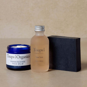 The Essential Facial Set by Toups and Co Organics, featuring a blue jar of frankincense tallow balm, a clear bottle of Clear Tonic, and a rectangular black bar of activated charcoal face soap, is displayed on a neutral background—perfect for achieving clear skin.