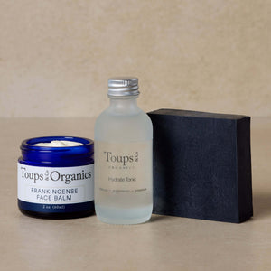 The Essential Facial Set from Toups and Co Organics—featuring a blue jar of Frankincense Tallow Balm, a frosted bottle of Hydrate Tonic, and an Activated Charcoal Face Soap bar—is artfully arranged on a neutral background. Each product is thoughtfully formulated to help reveal clear skin.