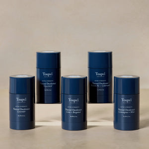 Five blue cylindrical containers of Toups and Co Organics Natural Extra-Strength Deodorant in assorted scents are arranged in a semi-circle on a neutral background.
