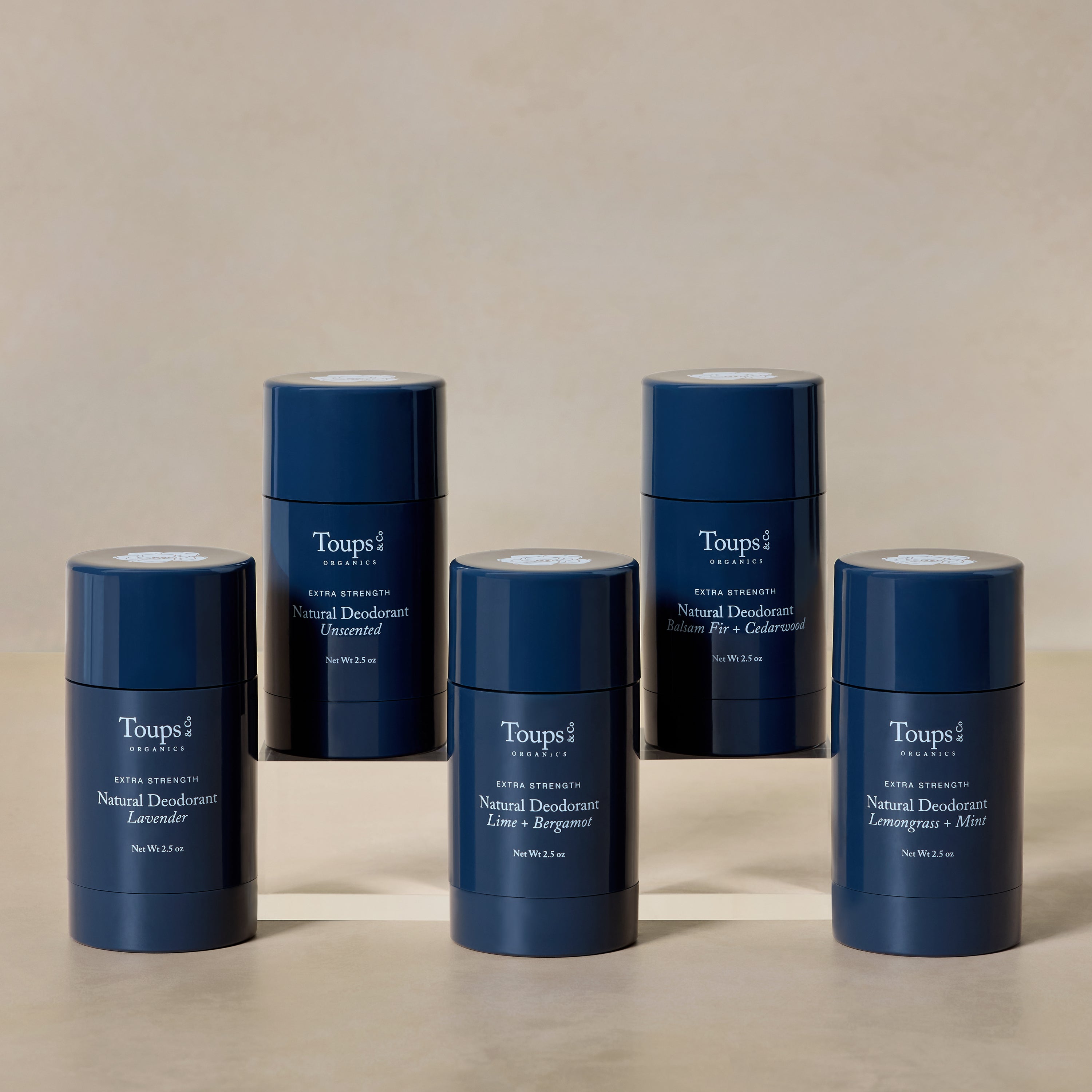Five blue cylindrical containers of Toups and Co Organics Natural Extra-Strength Deodorant in assorted scents are arranged in a semi-circle on a neutral background.
