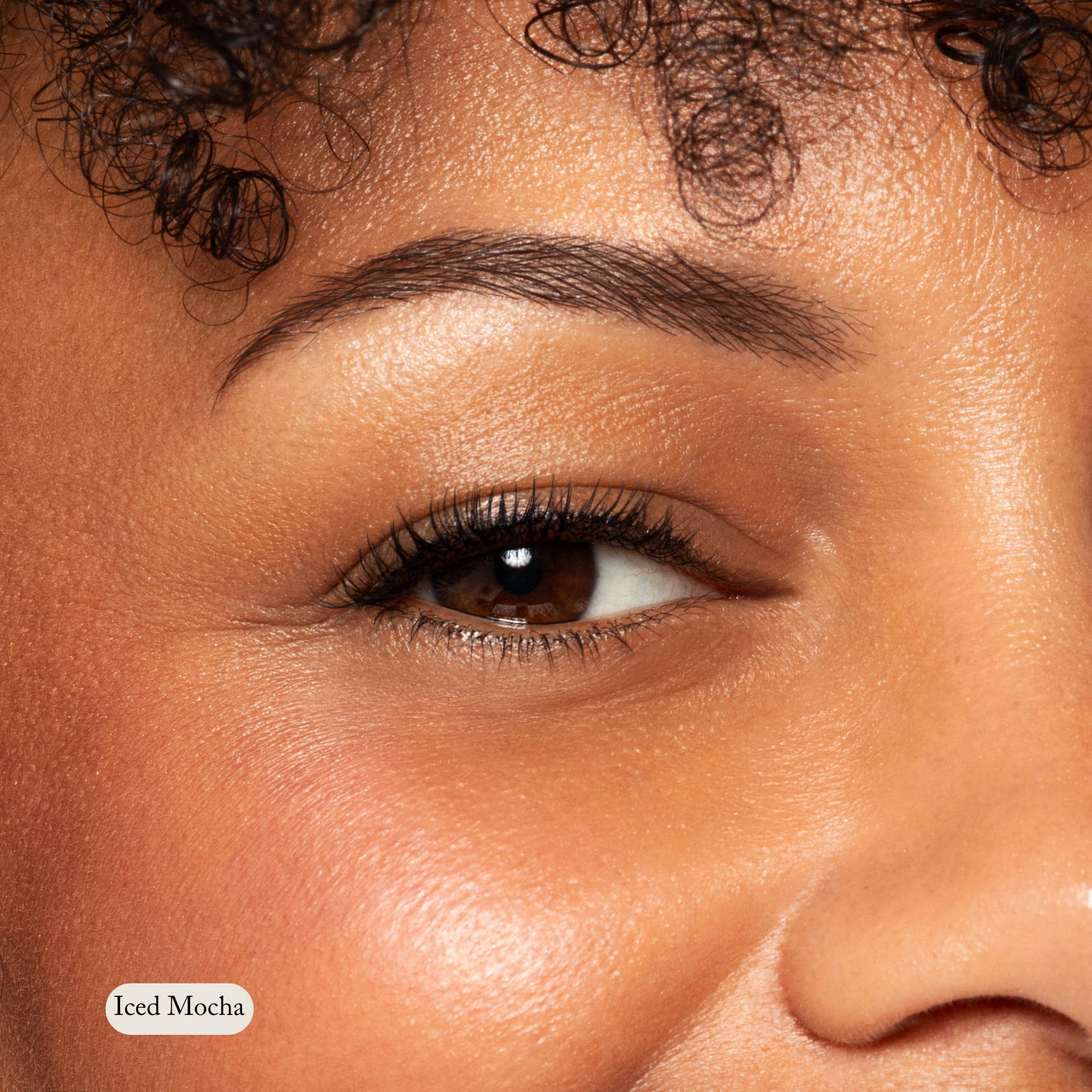 Close-up of an eye, eyebrow, and cheek with smooth brown skin and curly hair visible. Natural Eyeshadow Singles by Toups and Co Organics enhances the look. A label reading Iced Mocha appears in the bottom left corner.