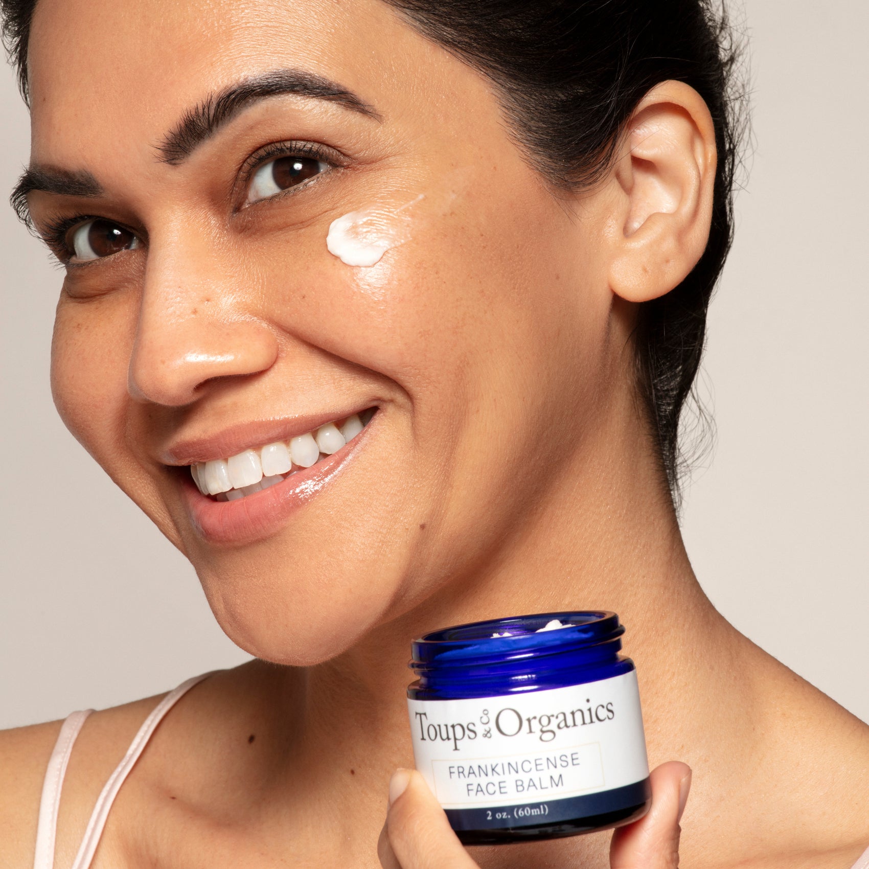 A woman smiles as she applies Toups and Co Organics Frankincense Face Balm, holding the blue jar of this nourishing, regenerative moisturizer near her cheek.