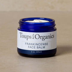 A blue jar labeled Toups and Co Organics Frankincense Face Balm, a nourishing tallow moisturizer, filled with white cream, sits on a beige surface.