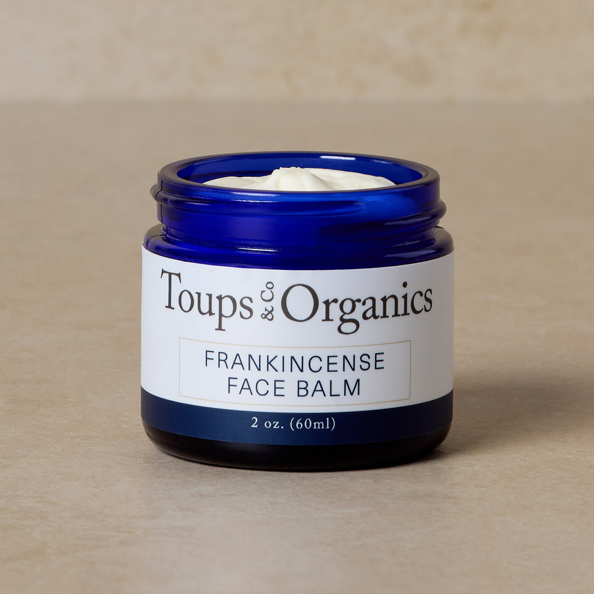 A 2 oz (60 ml) blue jar labeled Toups and Co Organics Frankincense Face Balm, a tallow-based moisturizer for reducing redness, rests on a neutral surface.