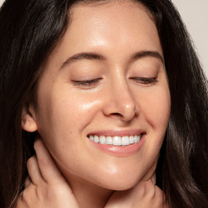A woman with long dark hair smiles with her eyes closed, showing radiant skin as she gently holds her neck—her glow enhanced by Glow Serum from Toups and Co Organics, infused with Helichrysum essential oil.