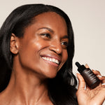 A woman smiles while holding a small dark glass dropper bottle of Toups and Co Organics Glow Serum, packed with vitamin-rich oils, close to her face.
