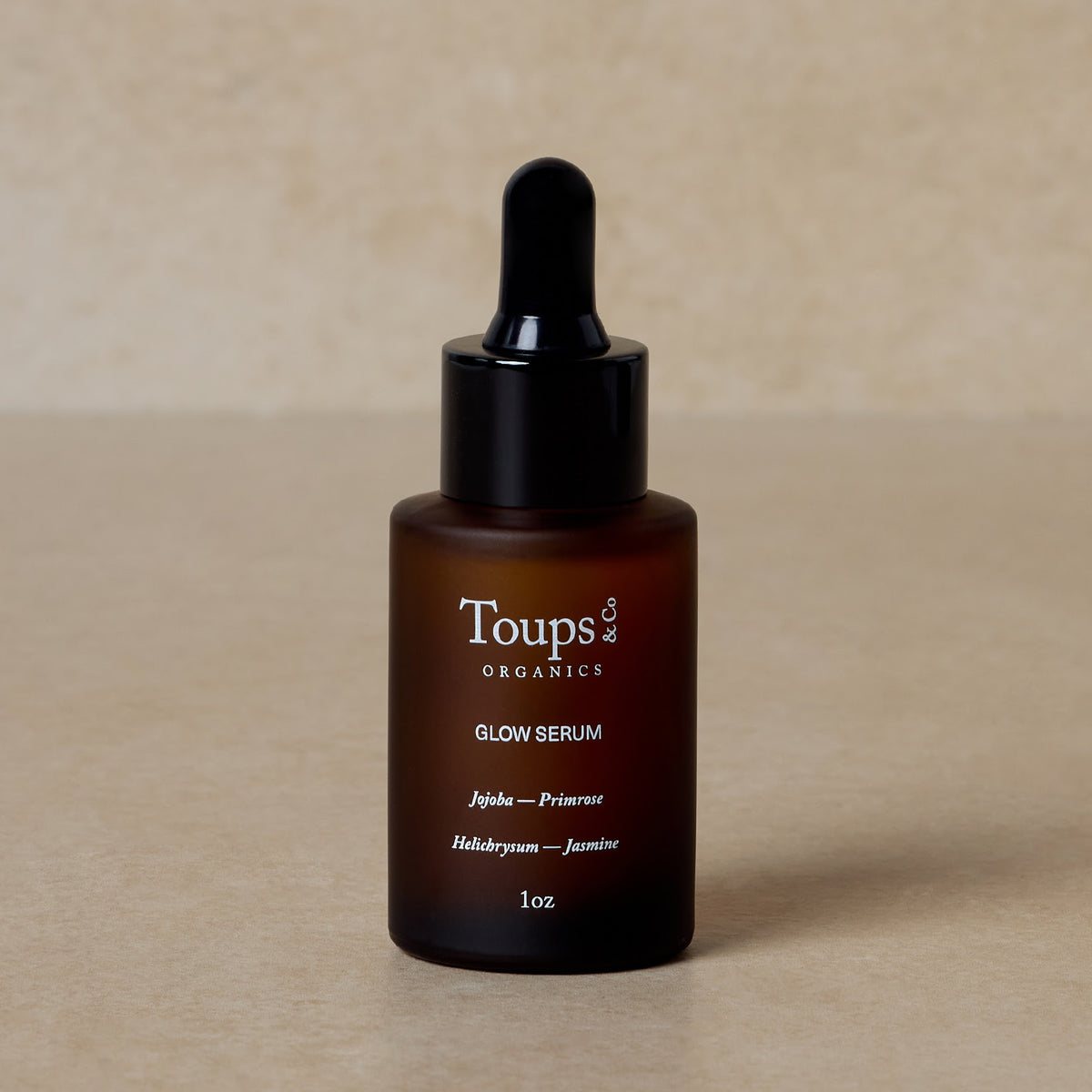 A brown glass dropper bottle of Toups and Co Organics Glow Serum, enriched with vitamin-rich oils for radiant skin, sits on a beige surface against a neutral background.