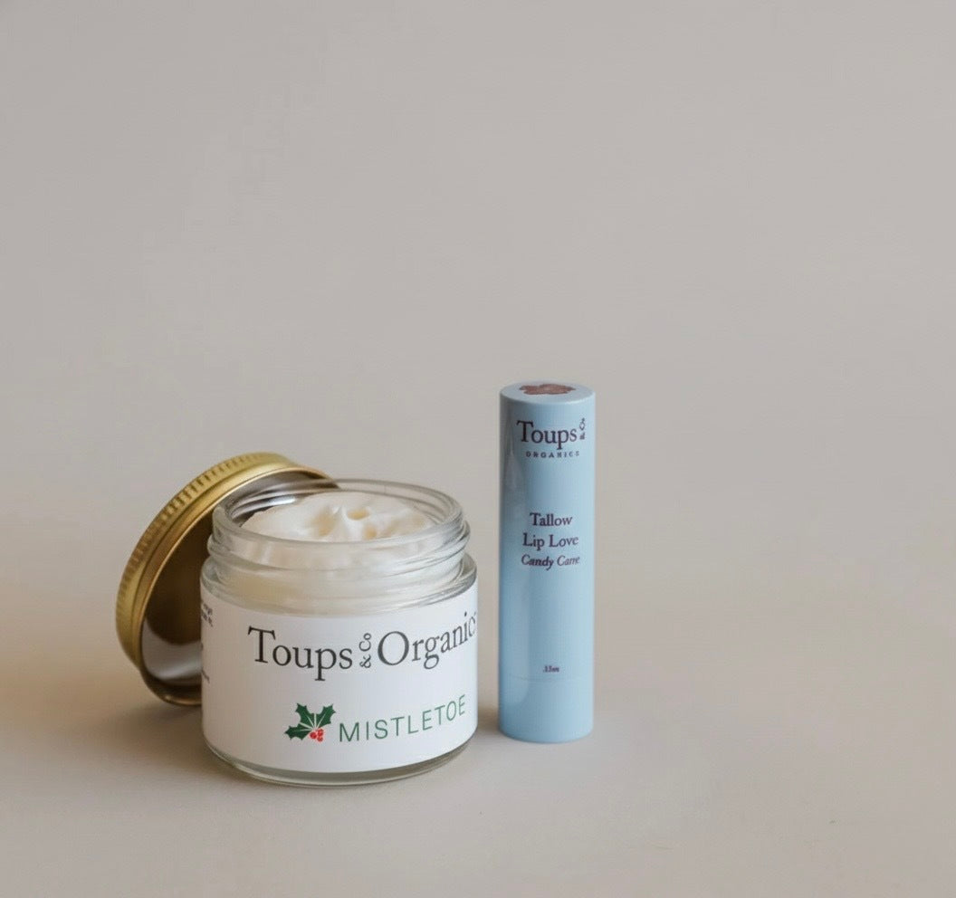 The Toups and Co Organics Merry and Bright Gift Set, featuring an open Mistletoe cream jar and a Tallow Lip Love balm in a light blue tube, is displayed on a plain background—natural skincare for the perfect holiday gift.