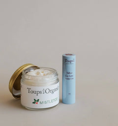 The Toups and Co Organics Merry and Bright Gift Set, featuring an open Mistletoe cream jar and a Tallow Lip Love balm in a light blue tube, is displayed on a plain background—natural skincare for the perfect holiday gift.