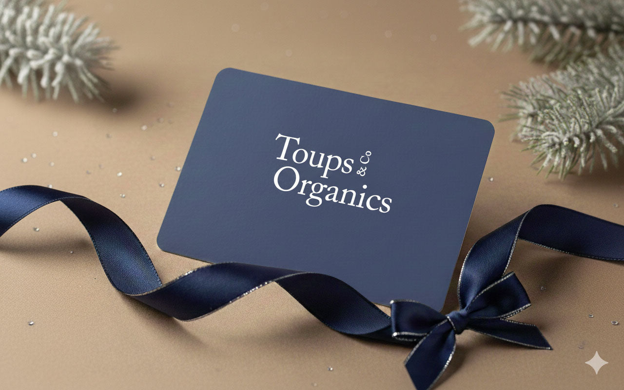 A dark blue Toups and Co Organics Gift Card rests on a tan surface with a matching ribbon and pine branches—perfect for gifting clean beauty and skin-loving products.