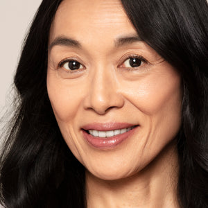 Close-up of a middle-aged woman with long black hair and clear skin, smiling gently as she showcases the brilliant pigmentation of Toups and Co Organics Highlighter while looking directly at the camera.