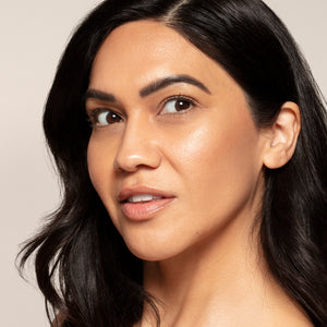 A woman with long dark hair and clear skin looks past the camera, her features enhanced by Toups and Co Organics Highlighter for brilliant all-natural pigmentation, set against a plain light background.