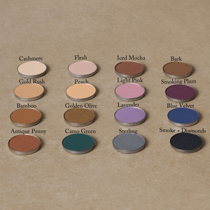 Sixteen Natural Eyeshadow Singles by Toups and Co Organics, featuring vibrant shades, are arranged in a grid on a beige surface, each labeled with its shade name.