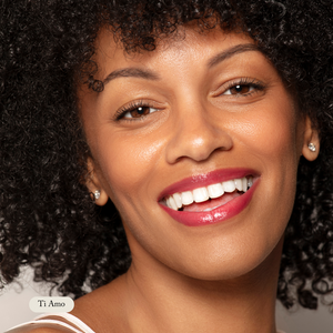 A woman with curly hair smiles at the camera, wearing stud earrings and moisturizing Organic Lip Gloss by Toups and Co Organics. Ti Amo appears in the lower left corner.