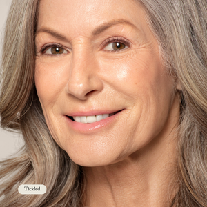 A middle-aged woman with long gray hair smiles softly at the camera; her lips shine with a smooth glow from Toups and Co Organics Organic Lip Gloss. A label in the corner reads Tickled.